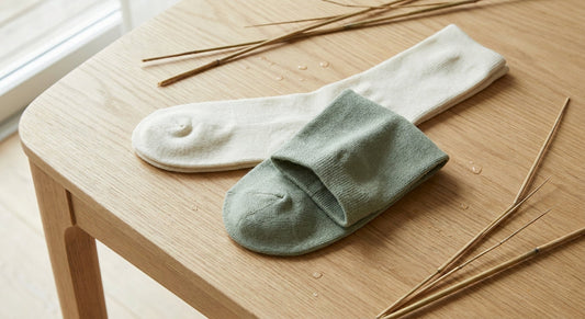 A close-up of bamboo fiber socks on a wood surface, one turned inside-out to show seamless toe construction, with bamboo stalks and water droplets nearby.