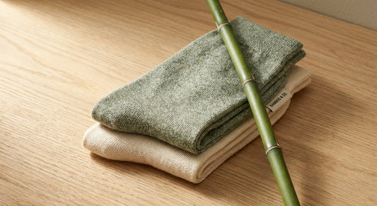 A close-up of neatly folded bamboo socks on a light wood surface beside a bamboo stalk, bathed in warm natural light.