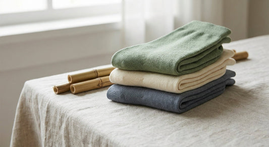 Folded bamboo socks in earthy tones arranged on a linen surface beside raw bamboo stalks in soft natural light.