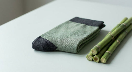 A pair of neatly folded bamboo socks resting next to fresh bamboo stalks on a clean light-grey surface, photographed in soft natural light.