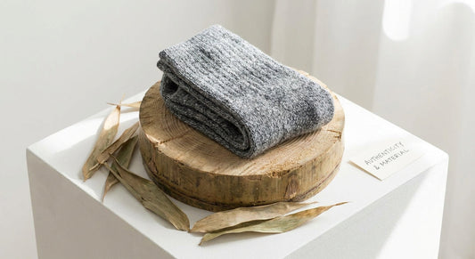 A pair of folded grey socks resting on a cross-section of raw bamboo stalk surrounded by bamboo leaves on a white surface.