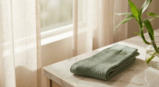 A pair of soft, neatly folded bamboo socks in a muted earthy tone resting on a marble surface beside a fresh bamboo sprig, lit with warm natural light.