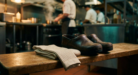 A pair of neatly folded bamboo socks resting beside work clogs on a wooden surface, with a softly blurred kitchen environment in the background.