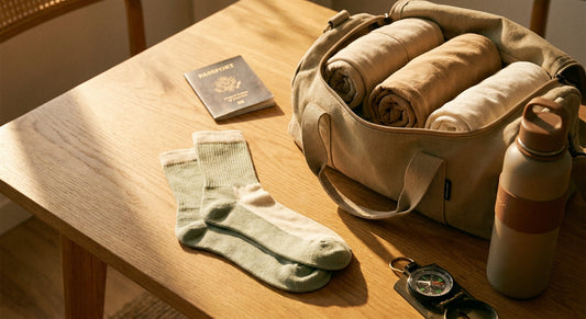 An open travel bag with neatly packed essentials including socks, a passport, and a water bottle arranged on a warm wooden surface in golden light.