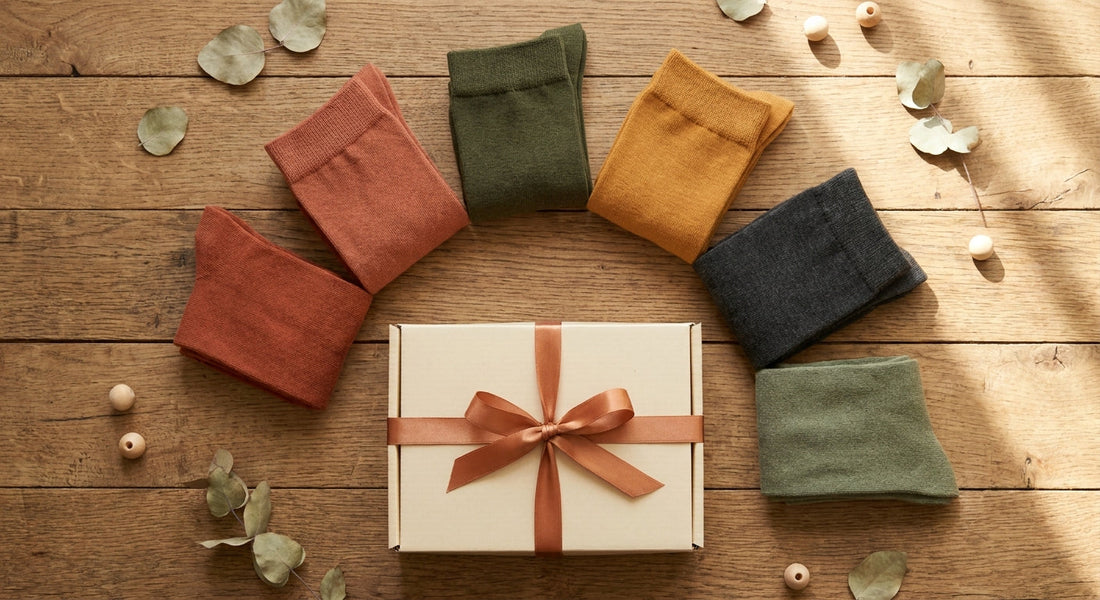 A styled flat lay of folded bamboo socks in earthy tones arranged around an open gift box with a ribbon on a warm wooden surface.
