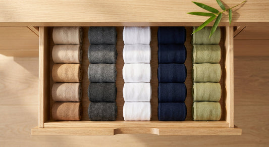 An overhead flat-lay of a neatly organized wooden sock drawer with five color-grouped rolls of bamboo socks arranged in a symmetrical grid under warm natural light.