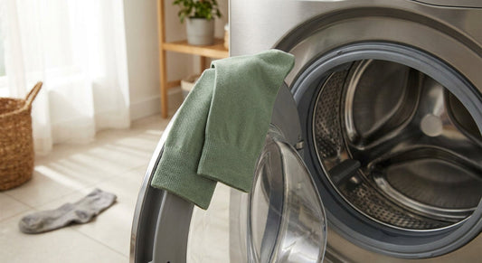 A pair of neatly folded sage green bamboo socks resting on an open washing machine drum, with a crumpled cotton sock discarded on the floor nearby in a bright, modern laundry room.